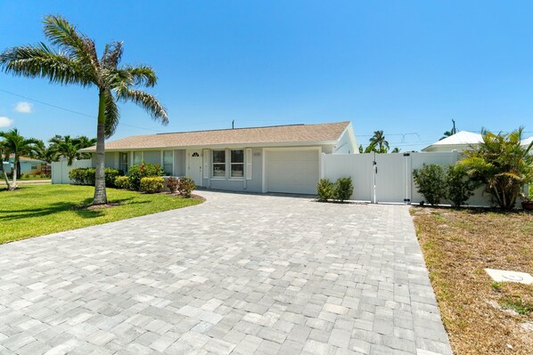 A single-story house with a paved driveway, a garage, and a small front yard with palm trees and shrubs.