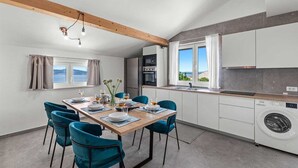 Una cocina moderna con horno empotrado y lavadora, un comedor con mesa de madera y sillas color turquesa, y vista al mar a través de amplias ventanas.