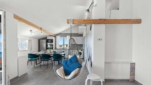 Cocina moderna con una silla colgante, comedor con sillas color turquesa y vista al mar a través de una ventana.