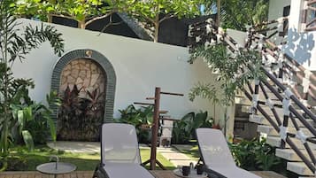Una terraza de madera con sillones y una pequeña mesa, rodeada de vegetación y un muro de piedra.