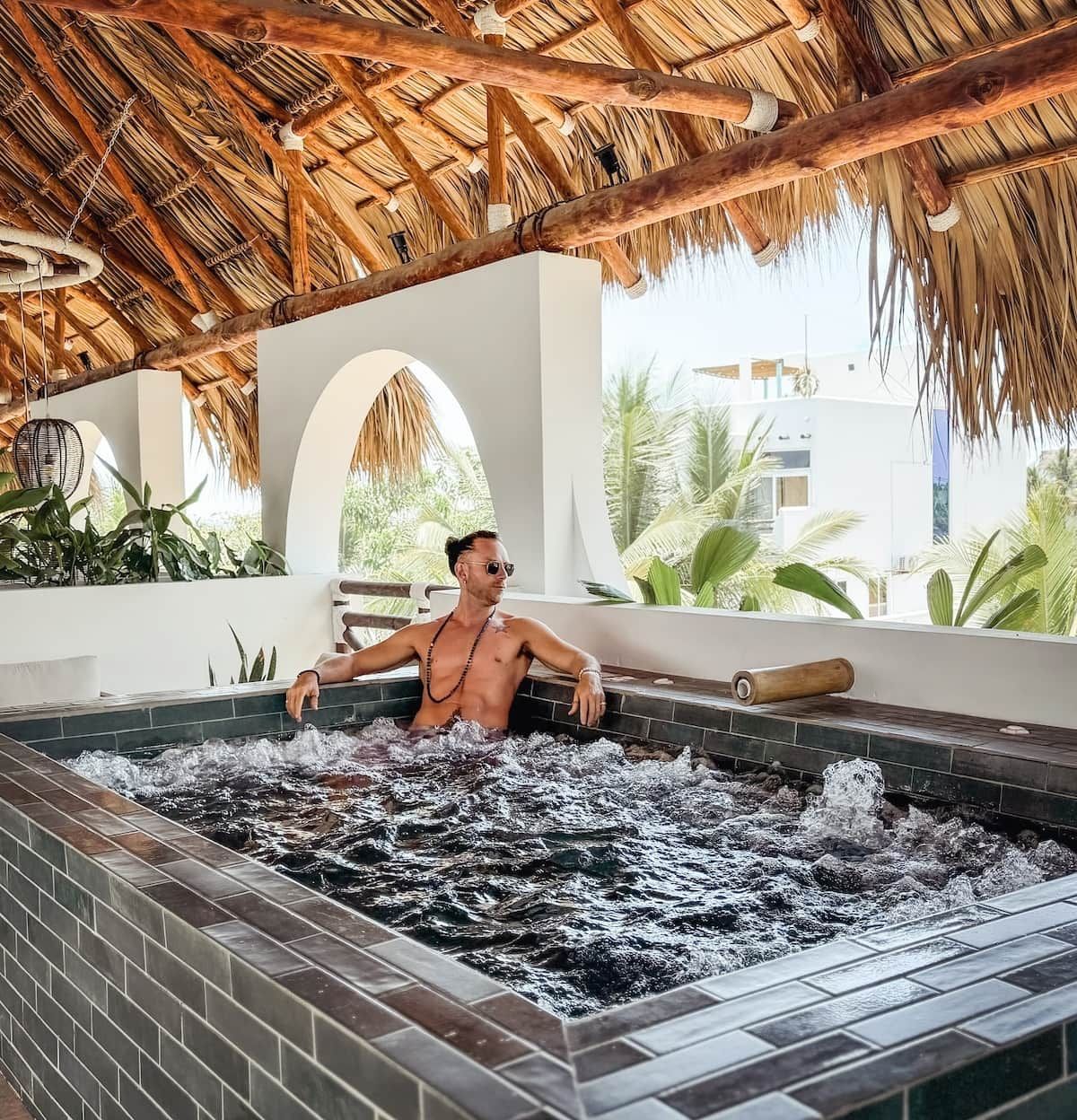 A man relaxing in a hot tub with a thatched roof overhead and a view of greenery outside.