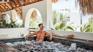 A man relaxing in a hot tub with a thatched roof overhead and a view of greenery outside.
