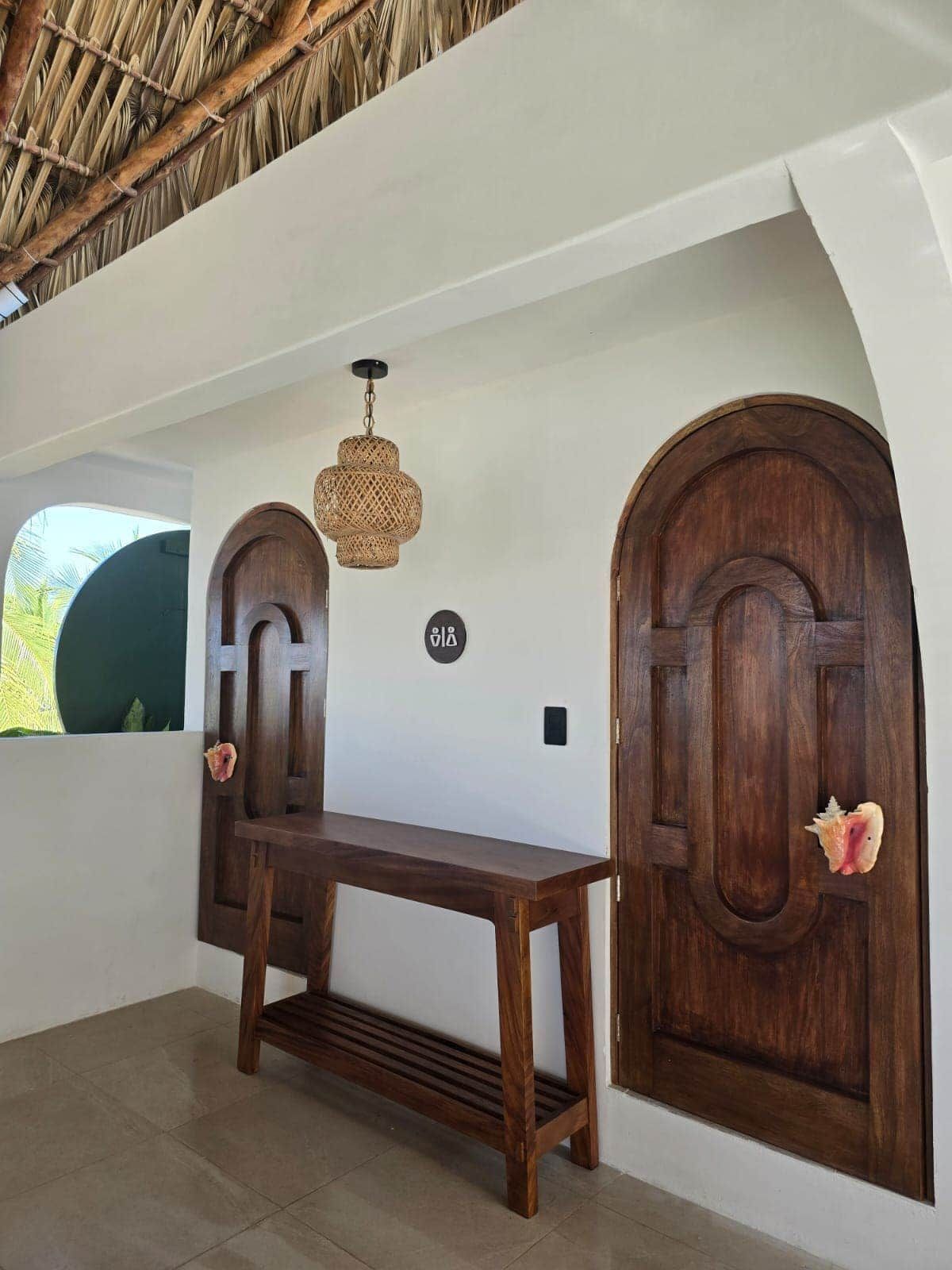Une pièce avec un banc en bois, deux portes en arc de cercle, un luminaire suspendu en fibres tressées et une petite fenêtre.