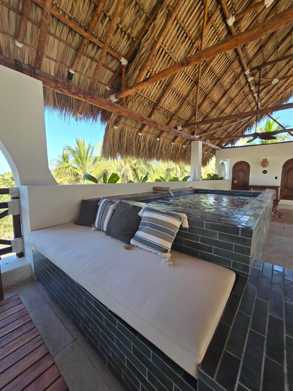 A hot tub area with a thatched roof, a cushioned bench, and a view of palm trees.