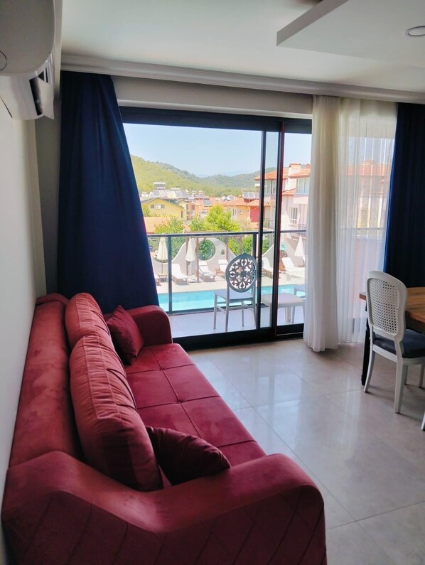 A red sofa in a room with a large window overlooking a pool and buildings. - Lina Suit (Fethiye)