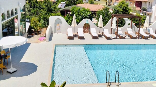 En swimmingpool med solstolar och parasoller, omgiven av byggnader och grönska.