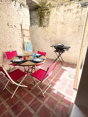 Uma área de refeições com uma mesa posta para quatro pessoas, cadeiras vermelhas, uma churrasqueira e uma escada encostada em uma parede texturizada.