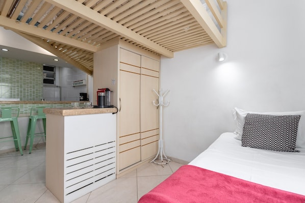 A bedroom with a bed, a red blanket, a white pillow, a wooden ceiling, and a kitchenette in the background.