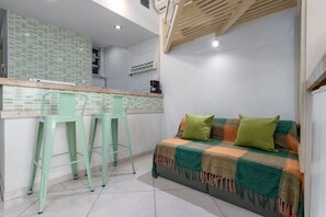 A kitchenette with a tiled backsplash, two green bar stools, a sofa with a checkered blanket, and a wooden ceiling.
