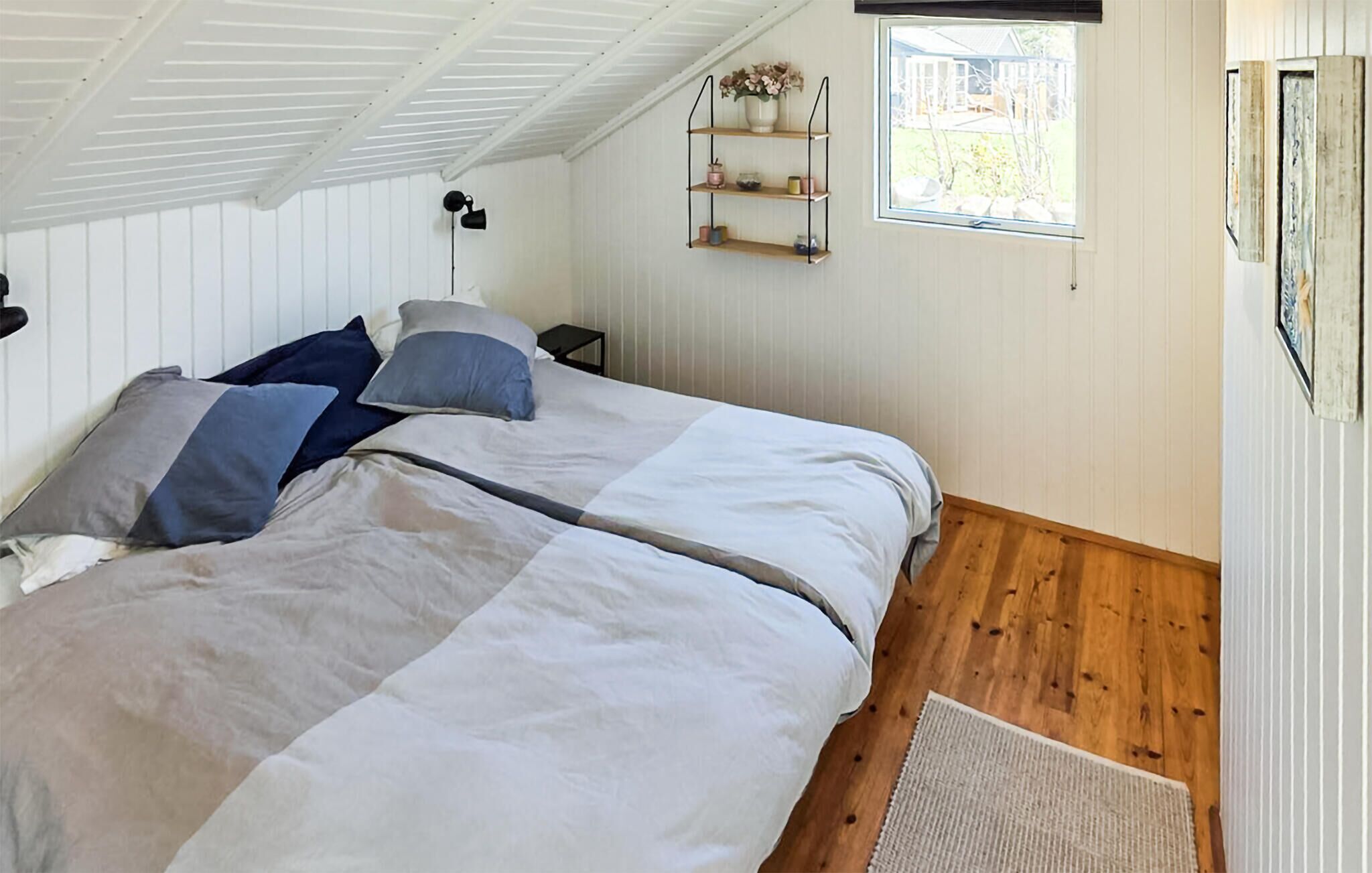 A bedroom with a bed, pillows, a nightstand, a shelf with items, a window, and a wooden floor.