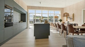 Une cuisine moderne dotée d’un îlot central, d’un coin repas avec des chaises en bois et de grandes fenêtres offrant une vue sur l’extérieur.
