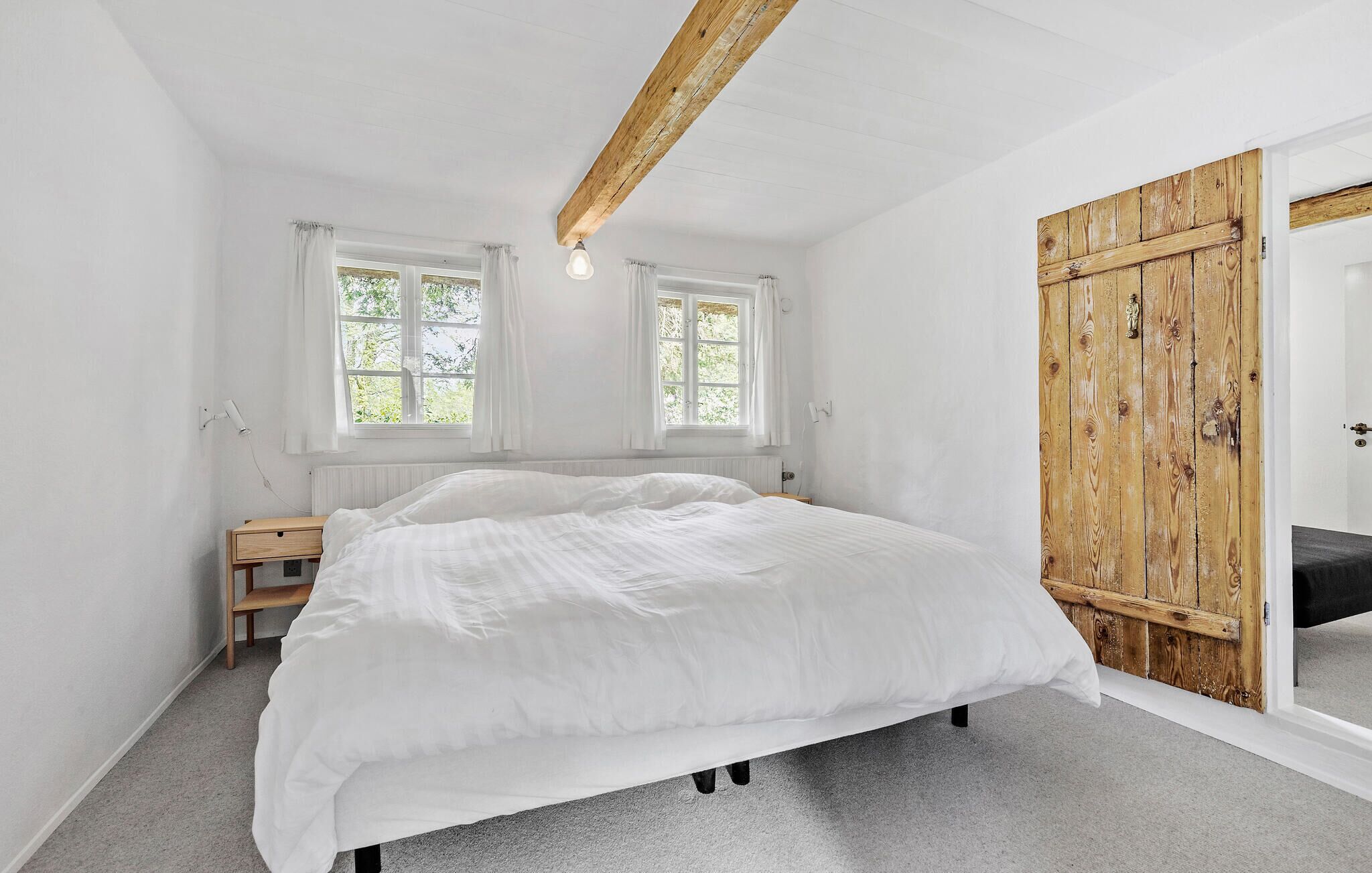 Ein Schlafzimmer mit einem großen Bett, zwei Fenstern, einer Holztür und einem kleinen Nachttisch.