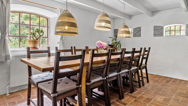 A dining area with a wooden table and chairs, two hanging wicker lamps, a potted plant, and a view of greenery through a window. - 5 bedroom cozy home in ørsted (Ørsted)