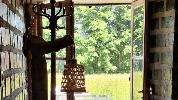 A rustic interior with a large wooden structure, a hanging lantern, and a view of a green field through an open door.