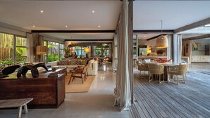 A spacious living area with a wooden floor, a dining area with wicker chairs, and a kitchen visible through an open doorway. - Your luxury home in the heart of Trancoso (Trancoso)