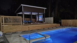 A swimming pool with a covered patio area, wooden deck, and a wooden fence.
