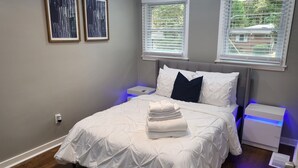 A bedroom with a bed, two bedside tables, a nightstand, and two windows with blinds.