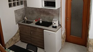 Cocina compacta con microondas, lavamanos y un refrigerador pequeño. Hay una puerta de madera con panel de vidrio y una ventana con diseño de cuadrícula en la pared.