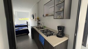 Une cuisine compacte équipée d’une cuisinière à gaz, d’une machine à café et d’un petit évier. La pièce adjacente contient un lit recouvert d’une couverture bleue.