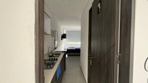 Un couloir étroit, avec un coin cuisine sur la gauche, un lit au fond et une porte en bois sombre sur la droite.