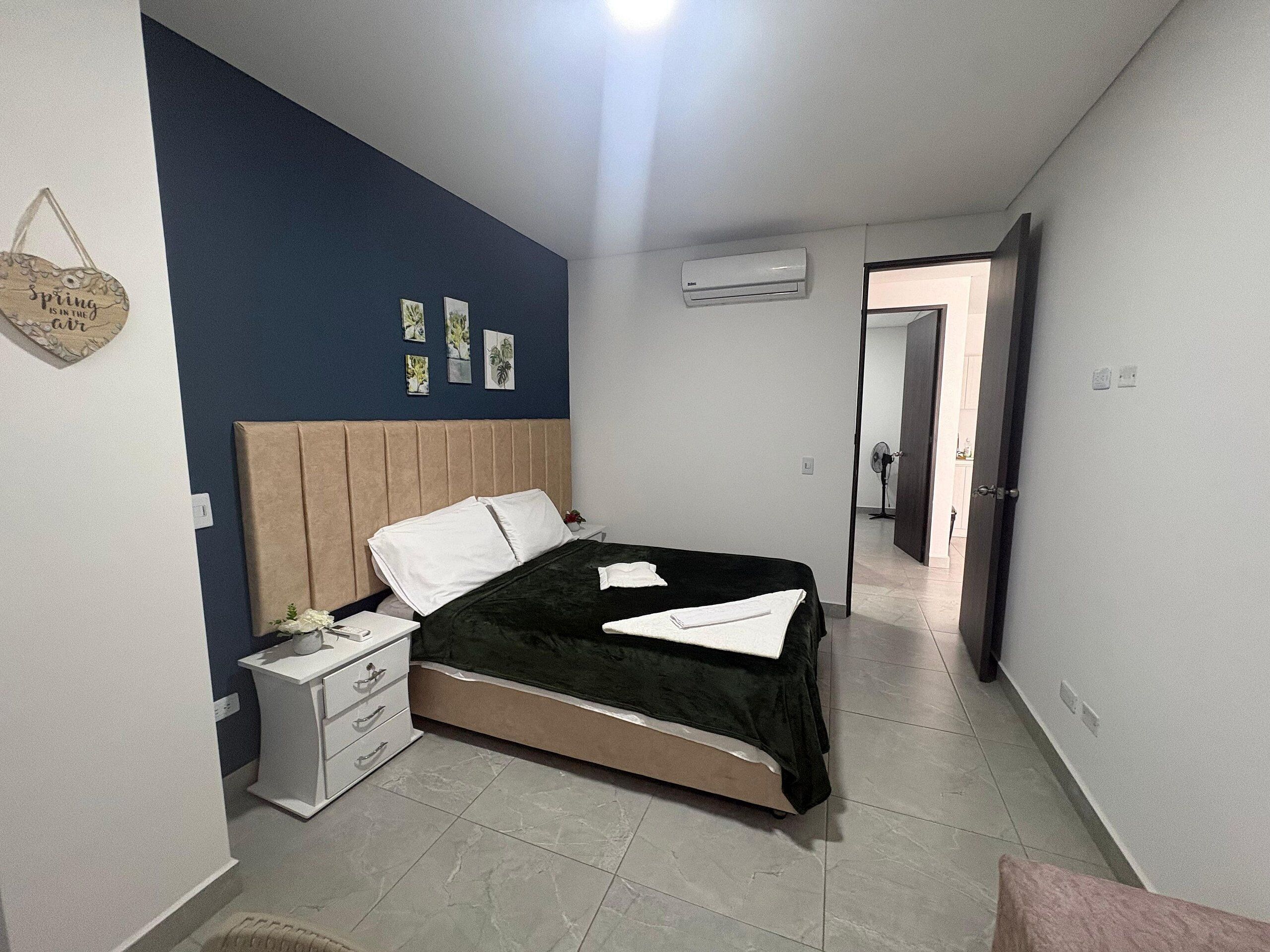 A bedroom with a bed, bedside tables, a nightstand, a wall-mounted air conditioner, and a door leading to another room.