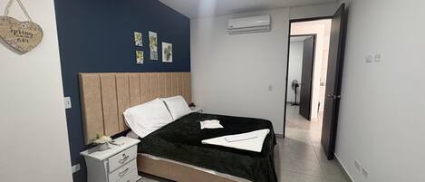 A bedroom with a bed, bedside tables, a nightstand, a wall-mounted air conditioner, and a door leading to another room.