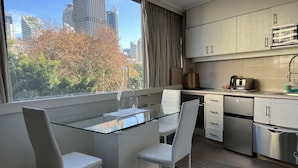 Una cucina moderna con un tavolo da pranzo in vetro, sedie bianche, elettrodomestici in acciaio inossidabile e vista sugli edifici della città.