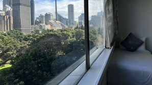 Ein Zimmer mit großem Fenster und Blick auf eine Skyline mit hohen Gebäuden und einem Turm, ein Bett mit Kissen sowie ein Fensterladen.