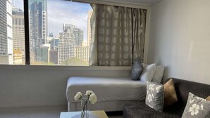 Ein modernes Zimmer mit einem Bett, einem Sofa und einem kleinen Tisch mit einer Blumenvase. Ein großes Fenster bietet Blick auf Stadtgebäude.