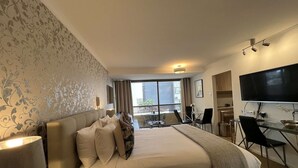 A hotel room with a large bed, a desk with a computer, a TV, and a balcony with outdoor seating.