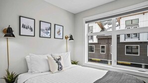 A bedroom with a bed, two bedside lamps, a window with a view of buildings, and three framed pictures on the wall.