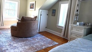 A bedroom with a curved chair, a dresser, a bed, a window with curtains, a door, and a framed picture on the wall.