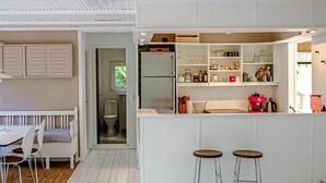 A modern kitchen with white cabinetry, a built-in oven, and a coffee machine. There is a dining area with a white table and chairs, and a small stool.