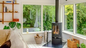 A cozy living room with a black wood-burning stove, large windows with a view of greenery, a bookshelf, and a comfortable sofa.