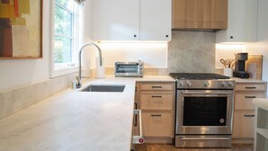 A modern kitchen with white cabinets, a stainless steel range, a built-in oven, a toaster, and a window with blinds. - Spanish-style garden bungalow! (Burbank)