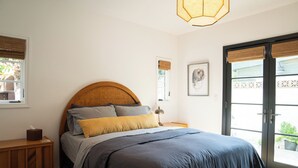 A bedroom with a bed, a bedside table, a lamp, a window with blinds, a door, and a door mat. - Spanish-style garden bungalow! (Burbank)