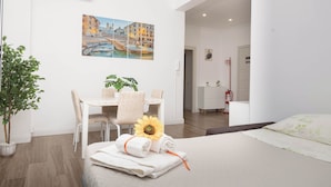 Una habitación de hotel con cama, un área de comedor con mesa y sillas, una planta en maceta, un cuadro con una vista urbana y un extintor.