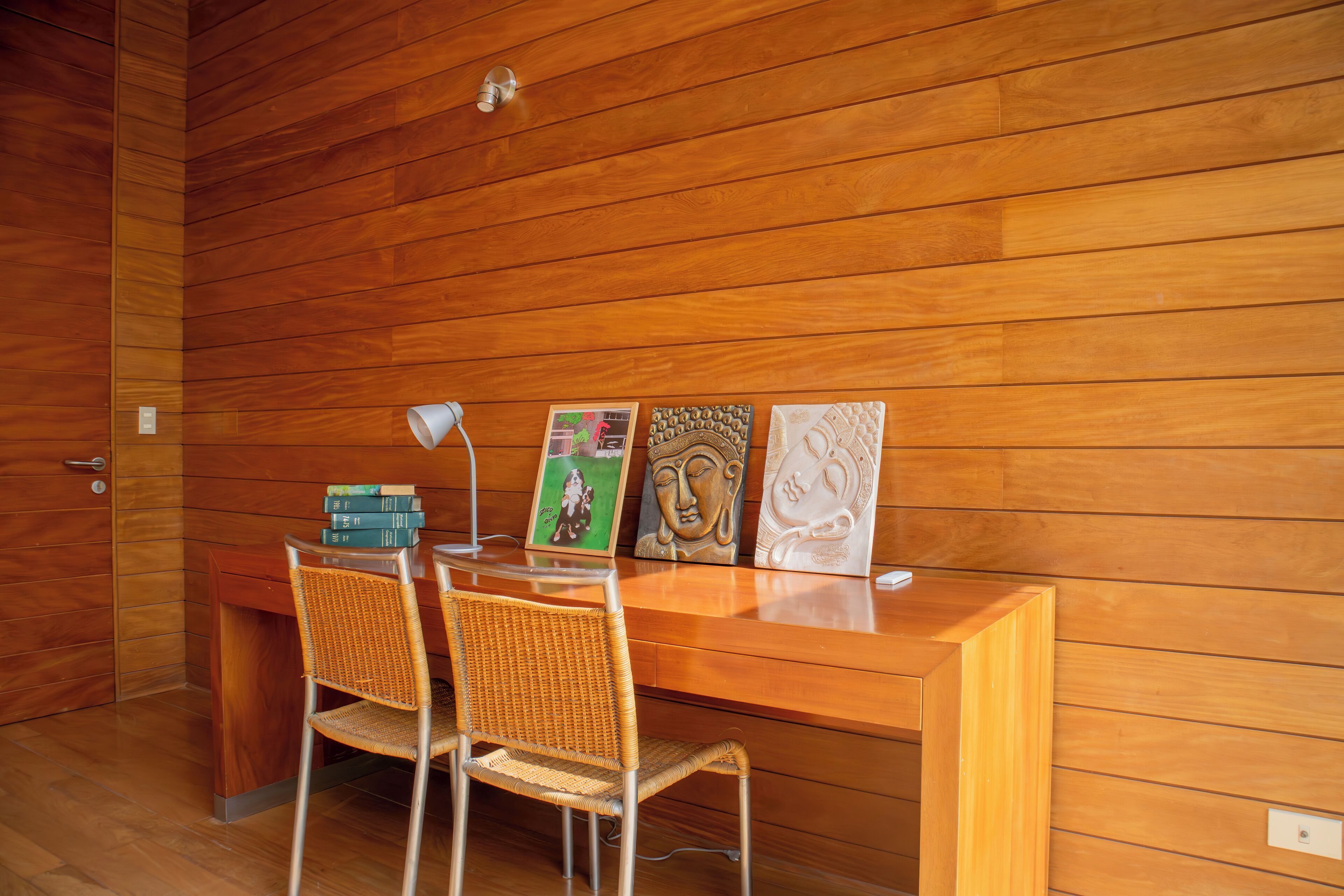 A wooden-paneled room with a desk, two wicker chairs, and three framed artworks.