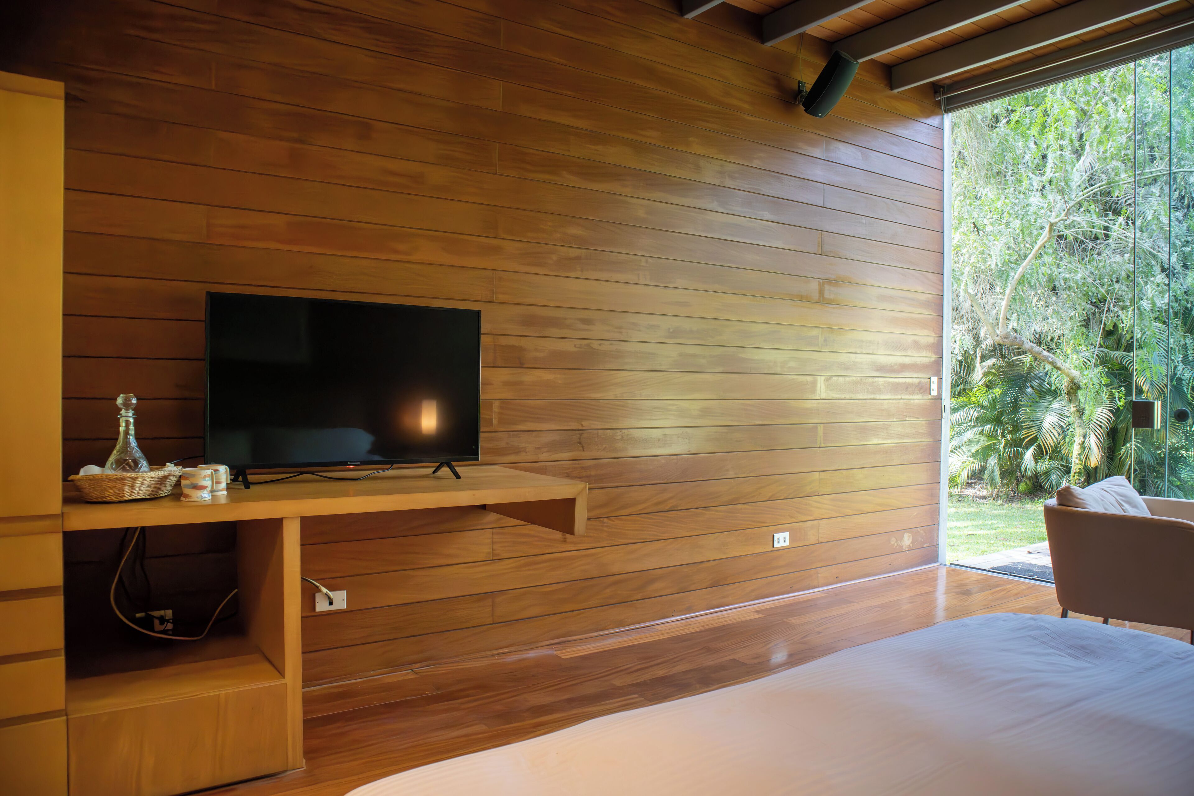 A modern room with a wooden wall, a flat-screen TV mounted on a wooden shelf, a bed, and a view of greenery outside.