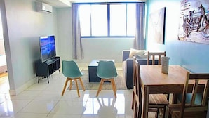 Una sala de estar moderna con un televisor de pantalla plana, sillas color turquesa, una mesa de comedor de madera y un ventanal con cortinas.