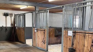 A stable with wooden stalls and metal gates, equipped with hay bales and grooming tools.