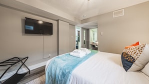 Una camera d'albergo con un letto, una TV montata a parete, una scrivania con una sedia e un bagno visibile attraverso una porta aperta.