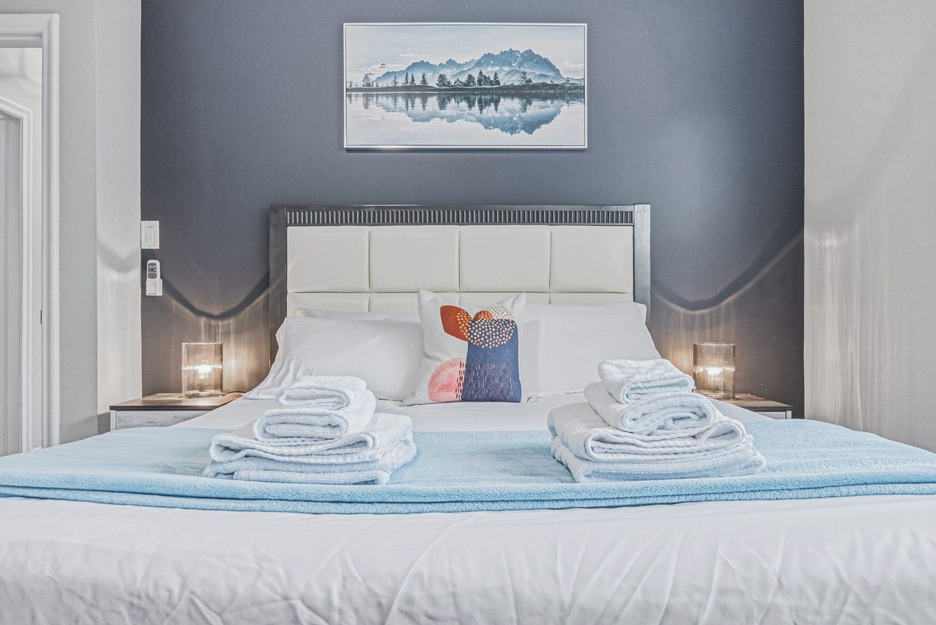 A neatly made bed with white linens and blue towels, a white headboard, and a framed picture of a mountain landscape on the wall.
