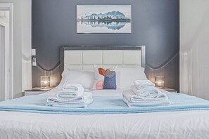 Un lit bien fait, avec du linge de lit blanc et des serviettes bleues, une tête de lit blanche et, au mur, un tableau représentant un paysage de montagne.
