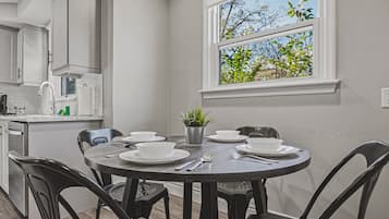 Un coin repas avec une table ronde dressée pour quatre personnes, entourée de chaises, d’une plante en pot et d’une fenêtre donnant sur des arbres.