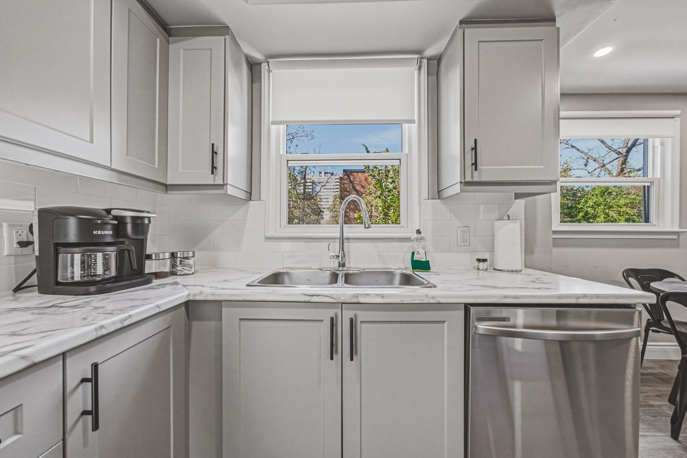 A modern kitchen with white cabinets, a built-in coffee maker, a sink, and a window with a view of trees and buildings.