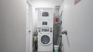 A laundry room with a front-loading washer and dryer, a small trash bin, and a shelf with cleaning supplies.