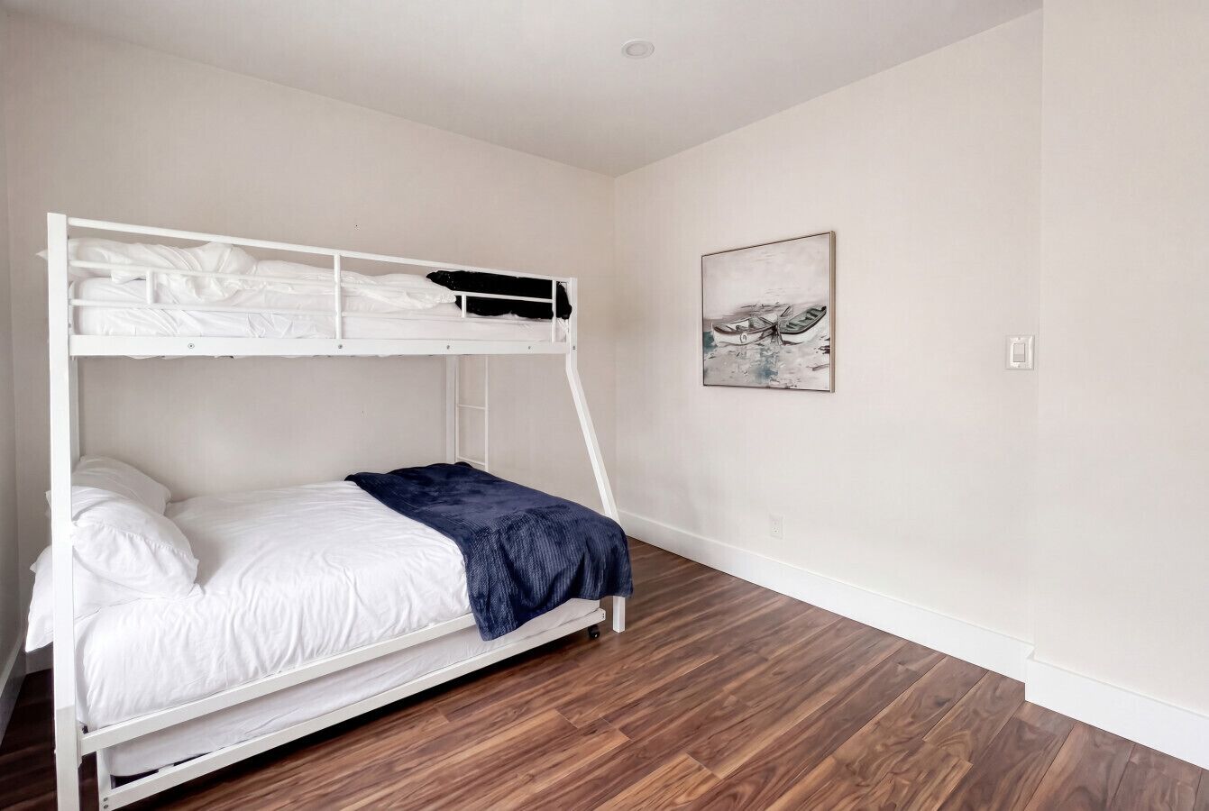 A bunk bed with white bedding and a blue blanket, a wooden floor, and a wall with a framed picture.