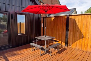 Une terrasse en bois avec une table et des bancs en métal, un parasol rouge et une clôture en bois.