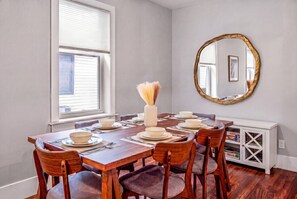 Een eetkamer met een houten tafel gedekt voor zes personen, een vaas met gedroogde bloemen, een ronde spiegel en een wit kastje met planken.